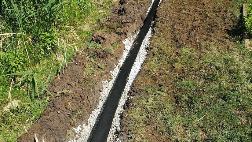 A drainage trench lined with black fabric and white gravel running diagonally across a lawn, with tall reeds on the left.